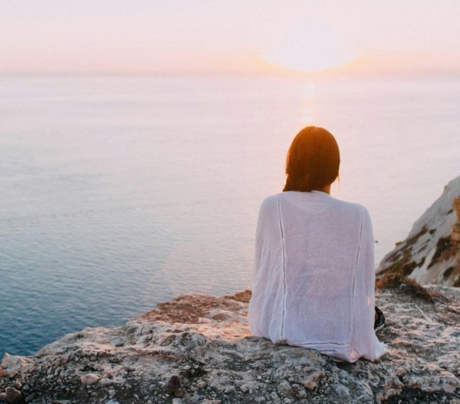 A serene view of a woman sitting on a rocky cliff, gazing at the ocean during sunset. Perfect for relaxation and travel themes.