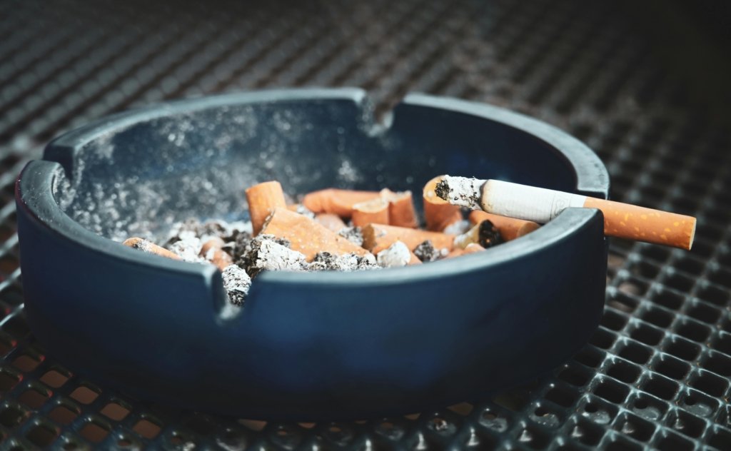 Close-up of cigarette butts and ashes in a blue ashtray with a smoldering cigarette, highlighting smoking habits.