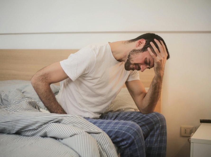 A young man in pajamas holding his head, sitting on a bed, appears to be experiencing a headache.