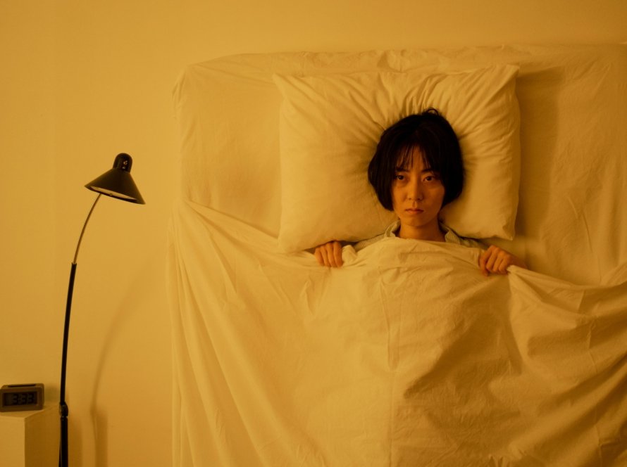 Woman lying under a blanket with a moody expression in a dimly-lit bedroom.